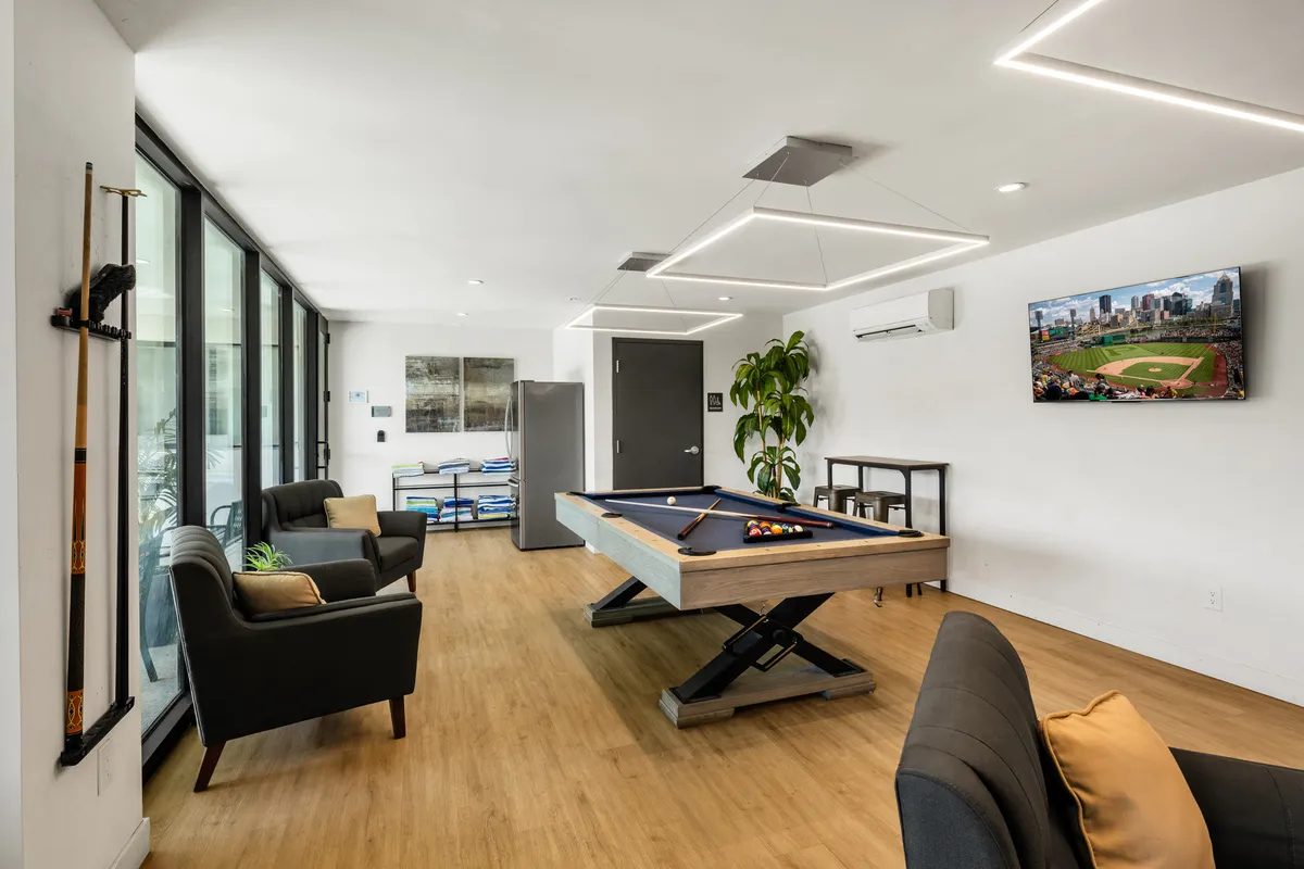 The image features a stylish clubhouse space with a pool table at the center, large windows flooding the room with natural light, a television screen for entertainment, and comfortable seating areas arranged for social interaction.