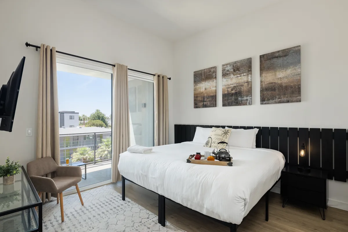A bright, inviting bedroom with a comfortable bed, soft linens, tasteful artwork, and a sliding glass door leading to a private balcony. Natural light fills the room, and there's a small table by the balcony for morning coffee or evening relaxation.