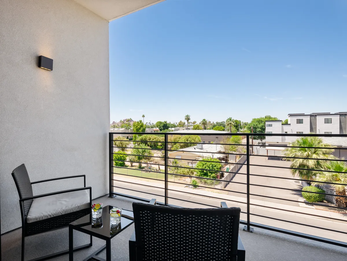 An exterior balcony scene featuring comfortable seating, a decorative tray of drinks, and a panoramic view of the surrounding neighborhood. The ambiance is calm and inviting, suggesting the perfect spot to gather with friends or start an evening out.