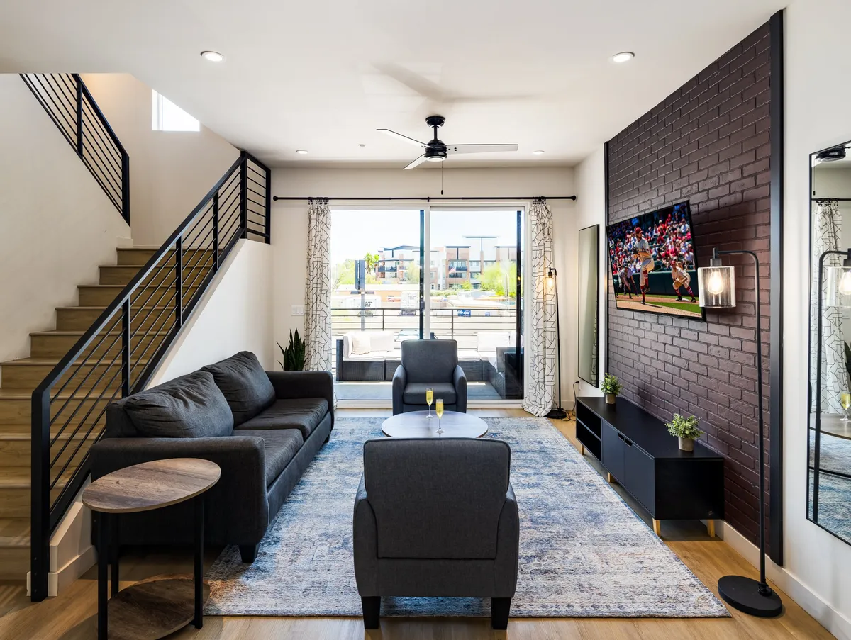 The image showcases a spacious, stylish living area with a plush sofa, modern coffee table, and a large TV, all framed by abundant natural light filtering through expansive windows. The open-concept design features a chic staircase leading to the second floor, creating a welcoming environment perfect for group gatherings and relaxation.