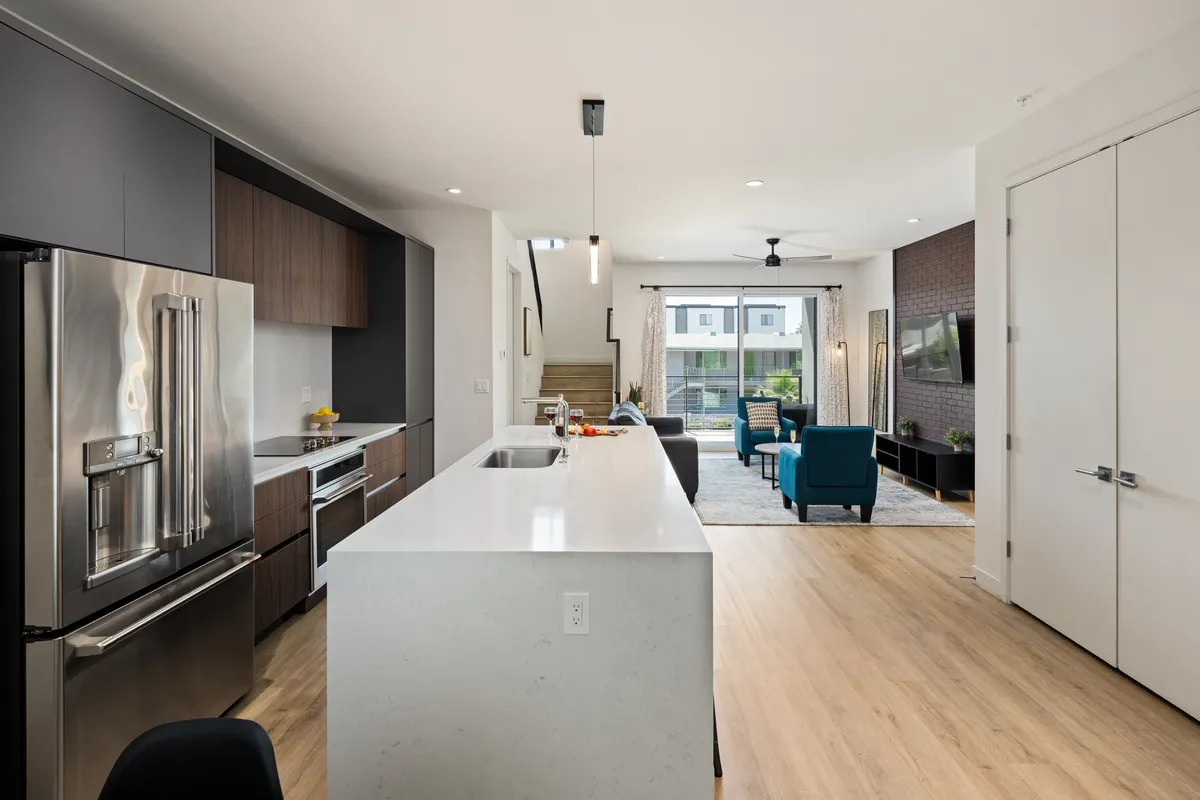 A bright, open-concept interior showing the kitchen island with bar seating, living area seating, and expansive windows that flood the space with natural light. The kitchen features modern appliances, and the layout showcases the seamless transition between kitchen, dining, and living areas.