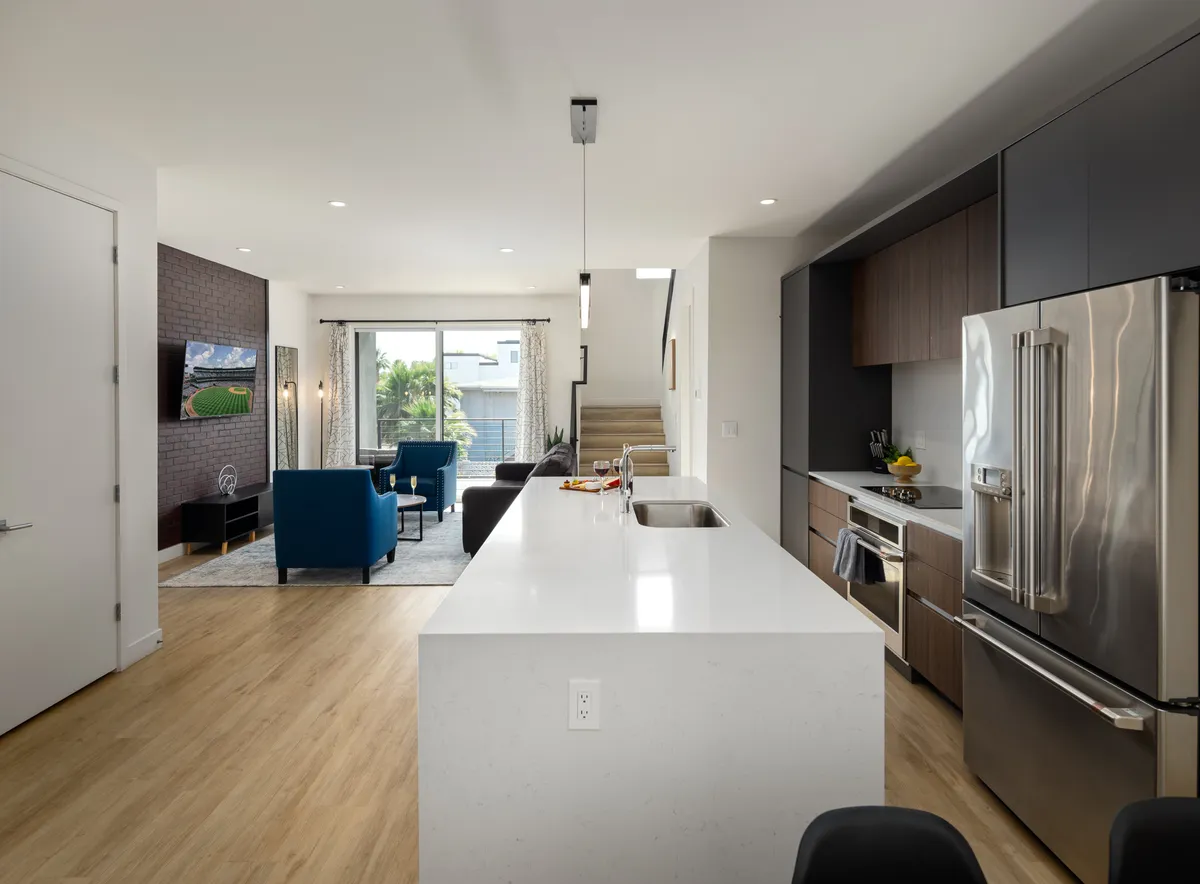 A stylish, modern open-concept living area with a kitchen island, living room seating, and expansive glass doors that lead directly to a furnished balcony. The layout seamlessly connects indoor relaxation with easy access to outdoor space.