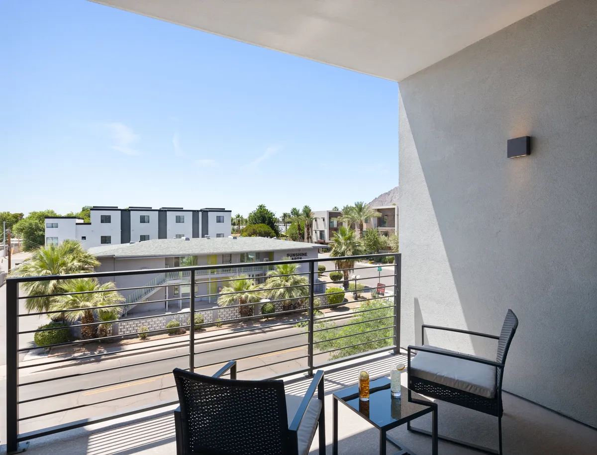 A tranquil balcony scene with comfortable outdoor seating, palm trees, and a view of surrounding buildings. The arrangement encourages group relaxation and small gatherings in a serene, sunlit atmosphere, making it ideal for after-hours networking or unwinding as a team.